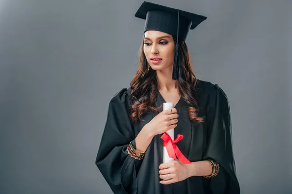 Индийская студентка в академическом платье и выпускной шляпе с дипломом, изолированный на сером — стоковое фото