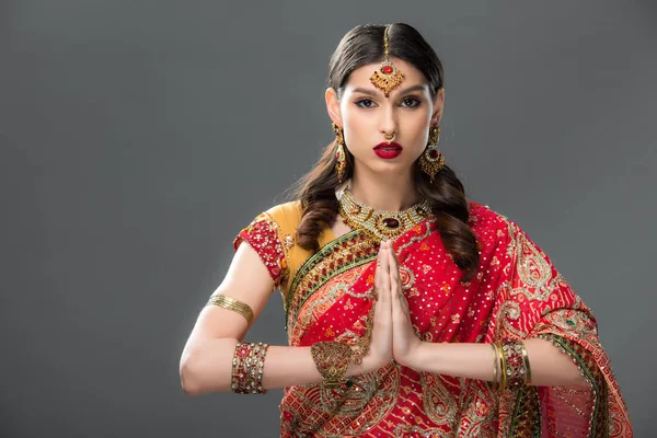 Индийская женщина в традиционной одежде и аксессуарах с мудрой намасте, изолированная на сером — стоковое фото