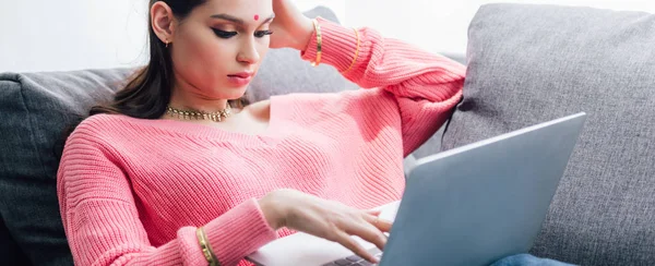 Attrayant indien femme avec bindi en utilisant ordinateur portable tout en étant couché sur canapé — Photo de stock