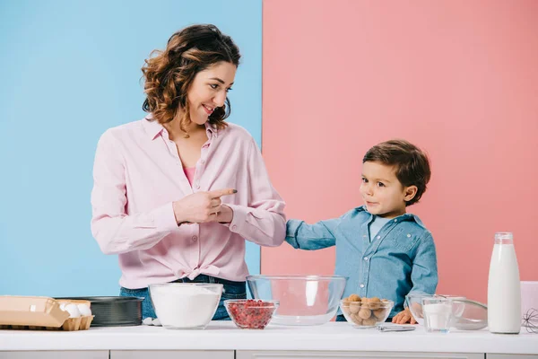 Sonriente madre con lindo hijo pequeño por mesa de cocina blanca con ingredientes para hornear sobre fondo bicolor - foto de stock