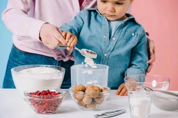 Attentive little boy with mother pouring flour into measuring cup together on bicolor background — Stock Photo