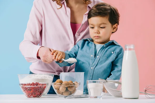 Милый маленький мальчик с матерью наливая муку в измерительную чашку на двухцветном фоне — стоковое фото