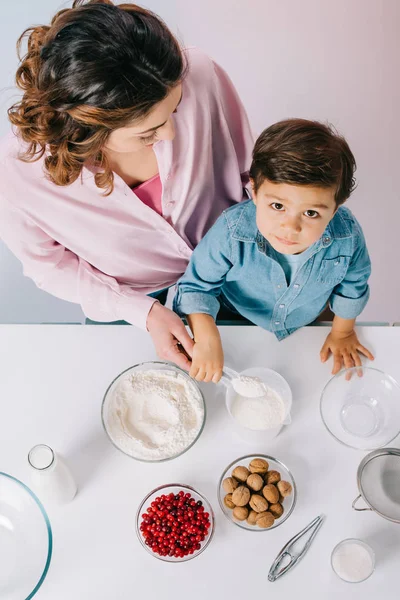 Вид сверху на симпатичного маленького мальчика и его маму, которые готовят вместе за белым кухонным столом на светлом фоне — стоковое фото