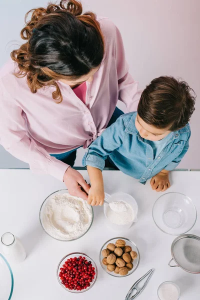 Top view of mother and little son pouring flour into measuring cup together on light background — Stock Photo
