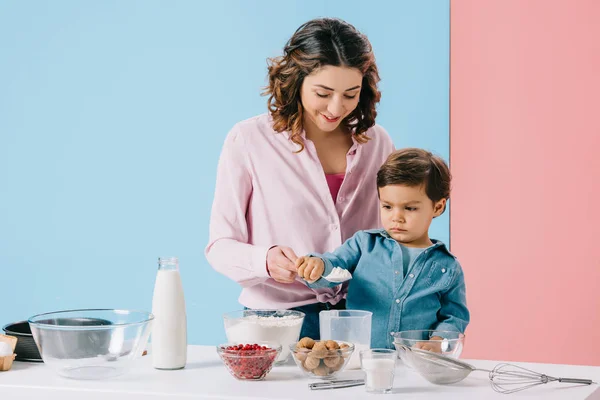 Улыбающаяся мама с милым маленьким сыном, смешивающим кулинарные ингредиенты на двухцветном фоне — стоковое фото