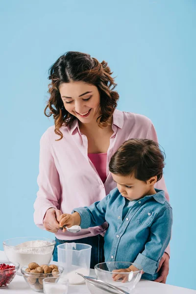Mãe feliz com adorável filho derramando farinha em copo de medição isolado em azul — Fotografia de Stock