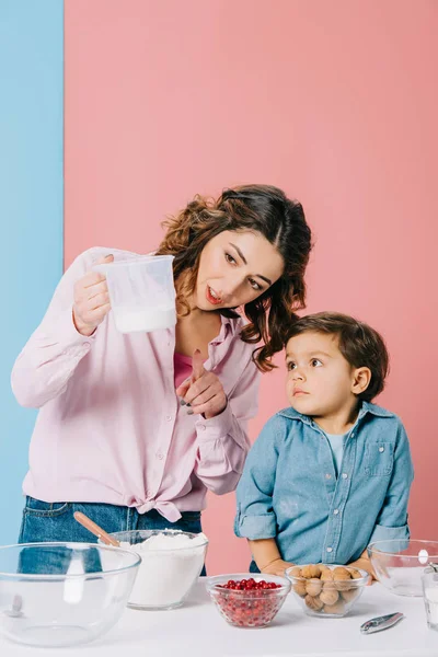 Красивая мать показывает номера на измерение чашки маленькому сыну на двухцветном фоне — стоковое фото