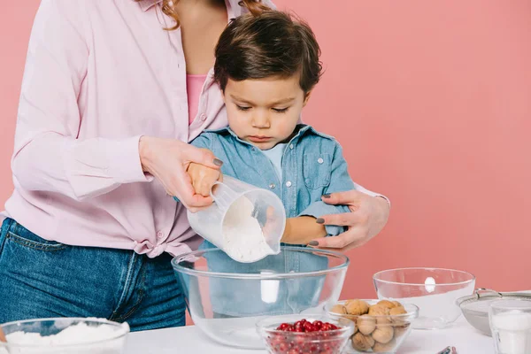 Vista parziale della donna che abbraccia il piccolo figlio mentre versa la farina nella ciotola insieme isolata sul rosa — Foto stock