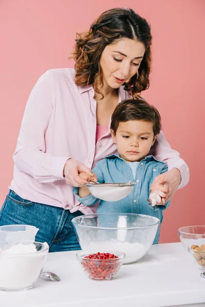 Bella madre con adorabile figlioletto setacciando farina con setaccio insieme isolato su rosa — Foto stock