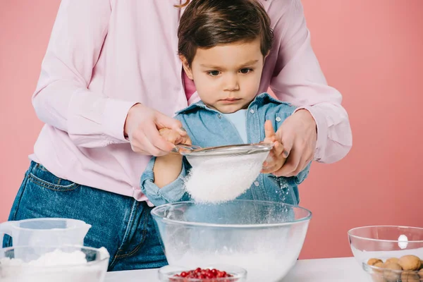 Serious little boy sifting flour together with mom isolated on pink — Stock Photo