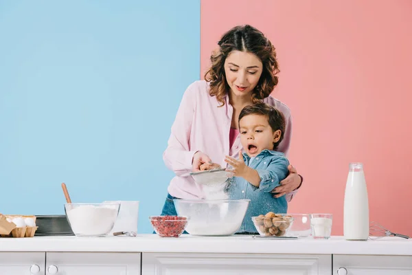 Madre con el pequeño hijo divertido tamizar harina para tazón sobre fondo bicolor - foto de stock