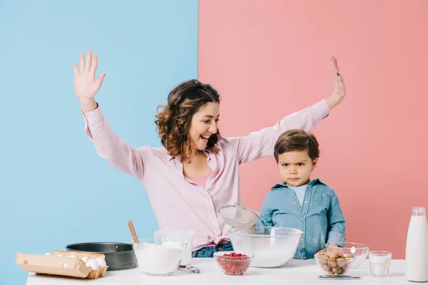 Улыбающаяся мать с поднятыми руками смотрит на серьезного маленького сына, держащего решето на двухцветном фоне — стоковое фото