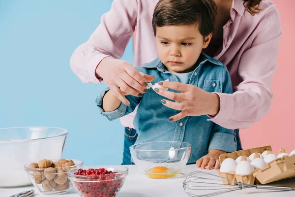 Netter kleiner Junge bricht Ei zusammen mit Mutter auf zweifarbigem Hintergrund — Stockfoto