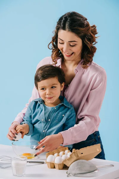 Feliz madre con sonriente pequeño hijo rompiendo huevo juntos aislado en azul - foto de stock