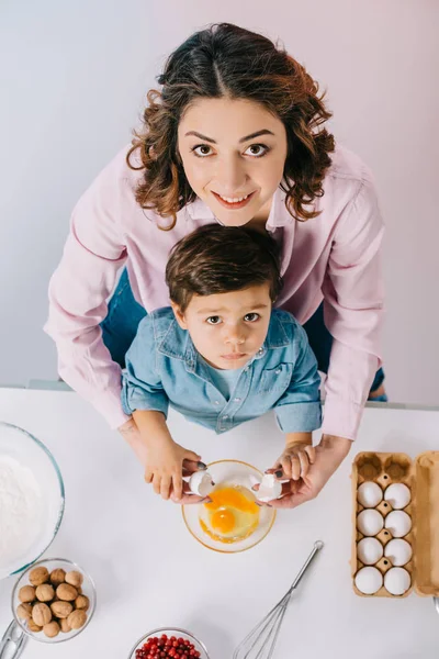 Верхний вид счастливой матери с очаровательным маленьким сыном разбивая яйца в миску и глядя на камеру на светлом фоне — стоковое фото
