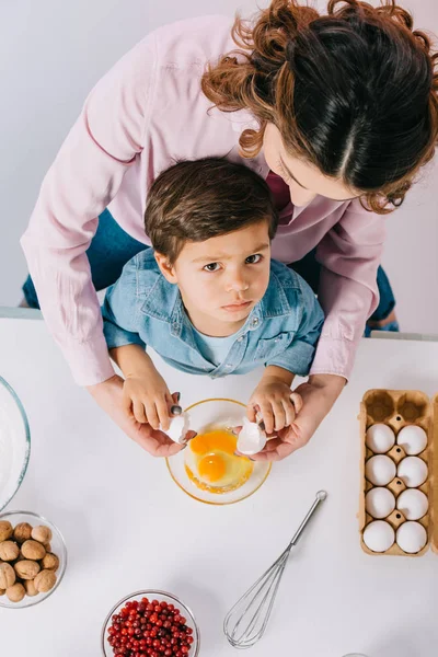 Верхний вид мамочки с серьезным маленьким сыном разбивая яйца в миску на светлом фоне — стоковое фото