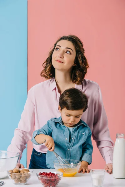 Madre alzando gli occhi e sorridendo mentre il piccolo figlio sbatteva le uova su sfondo bicolore — Foto stock