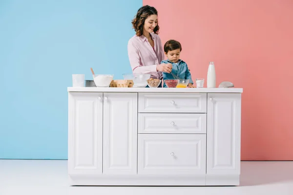 Glückliche Mutter mit entzückendem kleinen Sohn beim gemeinsamen Kochen am weißen Küchentisch auf zweifarbigem Hintergrund — Stockfoto