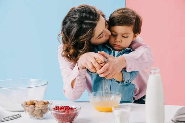 Mommy kissing adorable little son while whipping eggs with balloon whisk together on bicolor background — Stock Photo