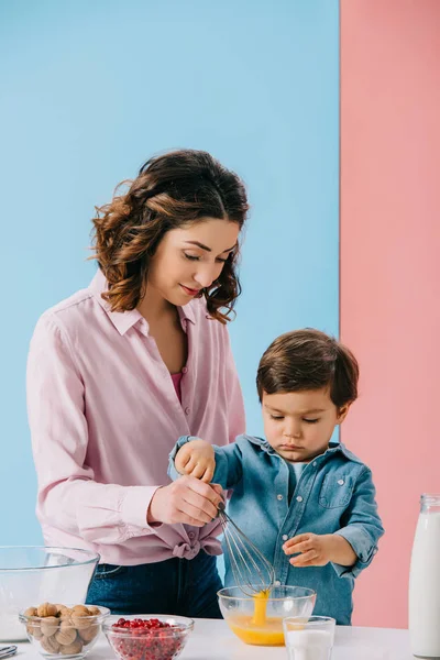 Madre ayudando a su pequeño hijo en la batida de huevos con batidor de globo sobre fondo bicolor - foto de stock