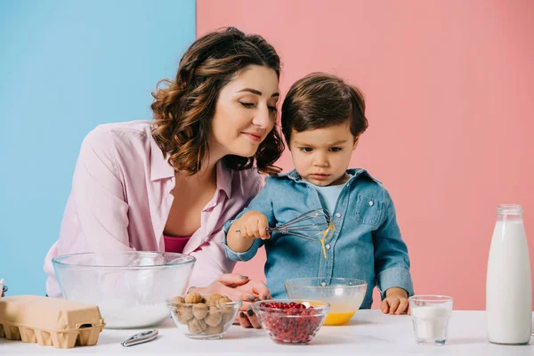 Feliz madre mirando lindo pequeño hijo batiendo huevos en bicolor fondo - foto de stock