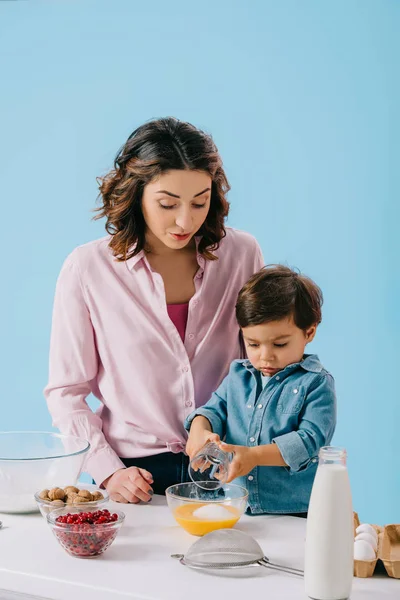 Cute little boy adding sugar to whipped eggs while mommy looking at him isolated on blue — Stock Photo