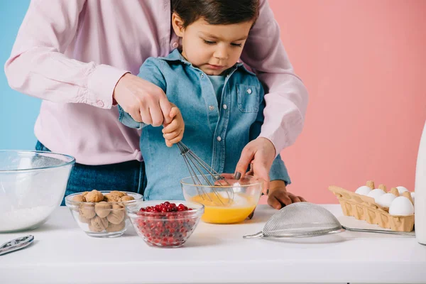 Милый маленький мальчик взбивая яйца с воздушным шаром в то время как мать помогает ему на двухцветном фоне — стоковое фото