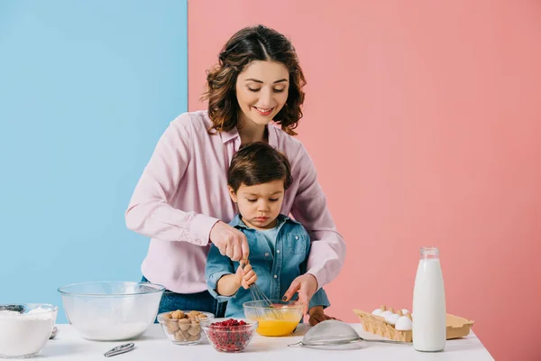 Sonriente madre ayudando a su pequeño hijo en la batida de huevos con globo batidor sobre fondo bicolor - foto de stock