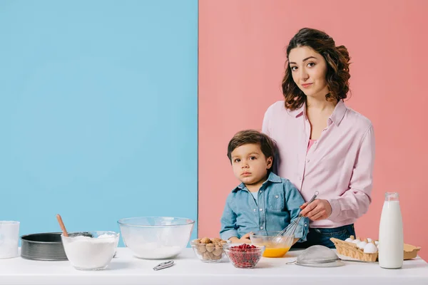 Mother with adorable son whipping eggs at white kitchen table with baking ingredients on bicolor background — Stock Photo