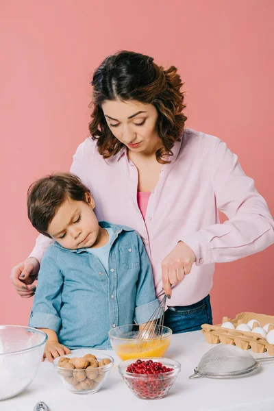 Netter kleiner Junge beobachtet aufmerksam seine Mutter beim Auspeitschen von Eiern isoliert auf rosa — Stockfoto