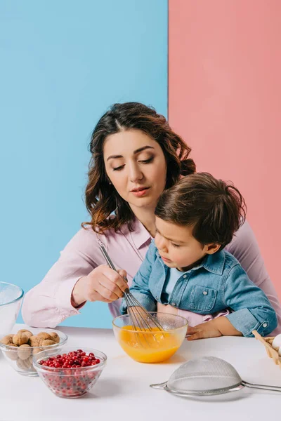 Мати сидить з маленьким сином за кухонним столом і збиває яйця в мисці на біколорному фоні — стокове фото