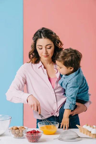 Mãe segurando pequeno filho enquanto chicoteando ovos no fundo bicolor — Fotografia de Stock