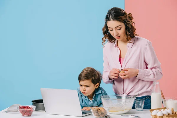 Netter kleiner Junge schaut auf Laptop-Display, während Mutter Walnuss auf zweifarbigem Hintergrund knackt — Stockfoto