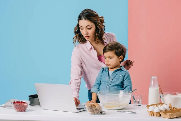 Madre usando escritorio mientras cocina junto con su hijo pequeño sobre fondo bicolor - foto de stock