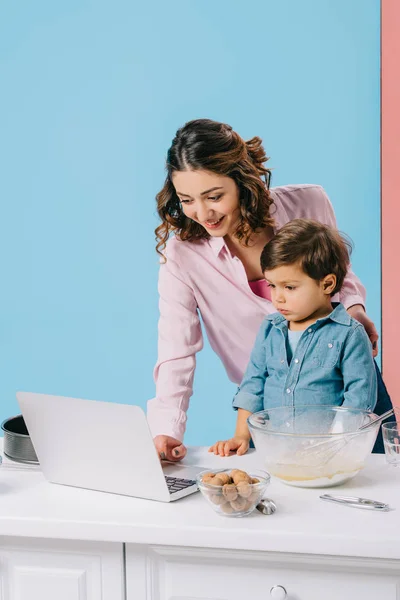 Mãe sorridente usando desktop enquanto cozinha junto com o pequeno filho no fundo bicolor — Fotografia de Stock