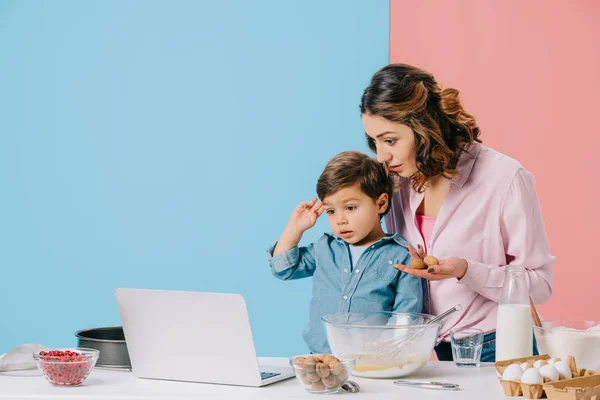 Madre usando escritorio mientras cocina junto con su hijo adorable sobre fondo bicolor - foto de stock