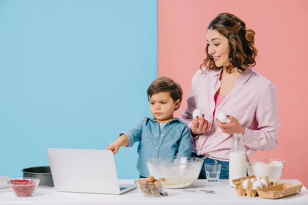 Menino apontando para a tela do laptop enquanto cozinha com a mãe no fundo bicolor — Fotografia de Stock