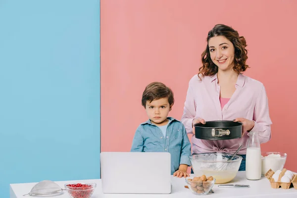 Mother holding baking form while cooking together with little son at kitchen table with products, cooking utensils and laptop on bicolor background — Stock Photo