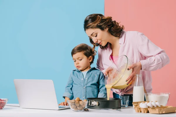 Madre verter la masa en forma de hornear, mientras que el pequeño hijo mirando la pantalla del ordenador portátil en fondo bicolor - foto de stock