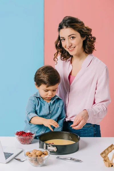 Lächelnde Mutter mit entzückendem Sohn, der Preiselbeeren zum Teig in Backform auf zweifarbigem Hintergrund hinzufügt — Stockfoto