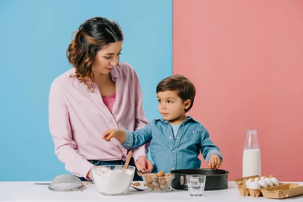 Madre feliz con adorable hijo pequeño cocinar juntos en la mesa de la cocina blanca sobre fondo bicolor - foto de stock