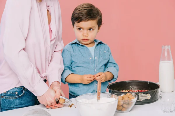 Lindo niño ayudando a la madre a agrietar nueces mientras cocinan juntos aislados en rosa - foto de stock