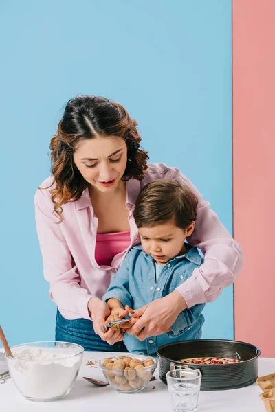 Mutter hilft kleinem Sohn beim Knacken von Walnüssen für Gebäck auf zweifarbigem Hintergrund — Stockfoto