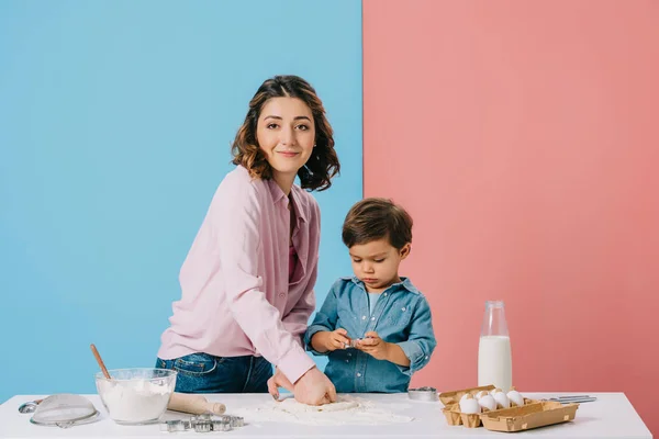 Lächelnde Mutter knetet Teig beim gemeinsamen Kochen mit kleinem Sohn auf zweifarbigem Hintergrund — Stockfoto