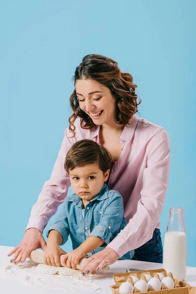 Mère heureuse avec adorable petit fils déroulant la pâte ensemble isolé sur bleu — Photo de stock