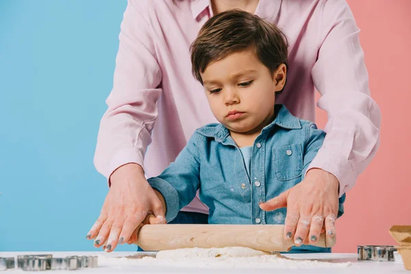 Милый маленький мальчик прокатывает тесто вместе с мамой на двухцветном фоне — стоковое фото