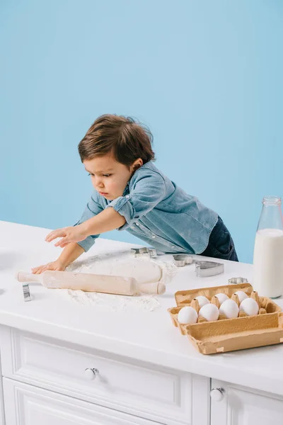 Mignon petit garçon déroulant la pâte avec rouleau à pâtisserie isolé sur bleu — Photo de stock