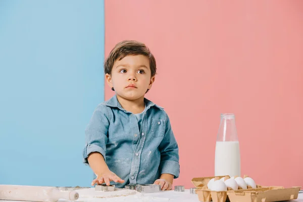 Маленька дитина вирізає фігури в тісті на білому кухонному столі на біколорному фоні — стокове фото
