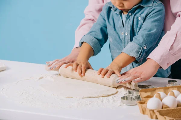 Vue partielle du petit garçon avec la mère déroulant la pâte ensemble isolé sur bleu — Photo de stock