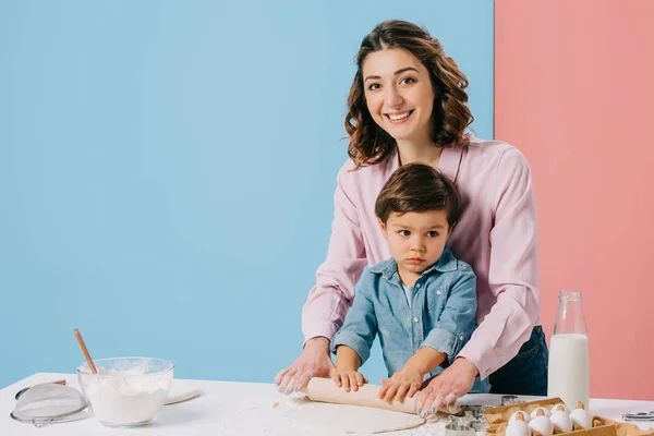 Lächelnde Mutter rollt Teig zusammen mit niedlichem kleinen Sohn aus und schaut in die Kamera auf zweifarbigem Hintergrund — Stockfoto